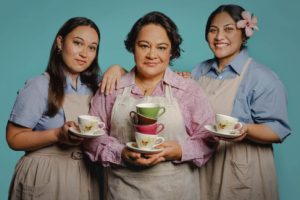 Three women stand together, they wear a work uniform and hold teacups